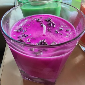 May include: A vibrant magenta candle in a clear, angled glass. The candle is topped with dried lavender and silver glitter. A white wick is centered in the candle. Other candles in green and orange are visible in the background.