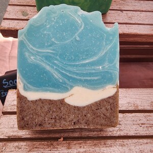 May include: A rectangular bar of soap featuring a layered design that mimics a coastal landscape. The top section is a marbled light blue, separated by a thin white band from the bottom section, which is a textured brown with dark speckles.