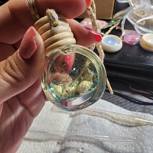 May include: A clear, round glass air freshener with a wooden cap and a rope for hanging. The interior contains small seashells, glitter, and a clear liquid. The air freshener is held by a person with pink nail polish.