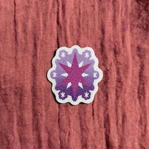 May include: A die-cut sticker featuring a starburst design in shades of purple and pink with white accents. The sticker has a glittery center and a white border, and is placed on a textured, reddish-brown fabric.