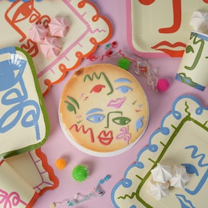 May include: A round cake decorated with colorful abstract face line art, surrounded by matching paper plates and cups featuring similar face designs, scattered white meringue cookies, and small colorful pom-poms on a light pink surface.