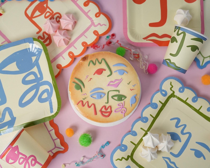 May include: A round cake decorated with colorful abstract face line art, surrounded by matching paper plates and cups featuring similar face designs, scattered white meringue cookies, and small colorful pom-poms on a light pink surface.