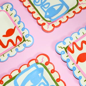 May include: A set of square paper plates with scalloped edges featuring a colorful abstract design. The plates display bold red, blue, and green line art patterns on a cream background, with some plates showing a stylized eye motif in blue.