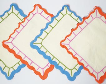May include: Four off-white square napkins with scalloped edges. Each napkin has a different color border: blue, orange, and pink. The interior of each napkin has a green border. The napkins are arranged diagonally.