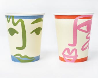 May include: Two paper cups with abstract faces drawn on them. The cup on the left has a green face with blue eyes and lips. The cup on the right has a pink face with orange eyes and lips.