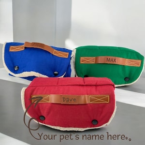 May include: Three personalized pet beds in blue, green, and red with brown leather handles. The beds have a white sherpa lining and are perfect for small dogs or cats. The beds are personalized with the names "Lila", "Max", and "Dave".
