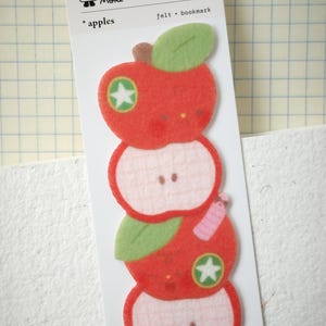 Peut inclure: Un marque-page en feutrine présentant une pile verticale de quatre formes de pommes sur une carte de support blanche. Le motif comprend deux pommes rouges entières avec des feuilles vertes et des étoiles, alternant avec deux moitiés de pommes.