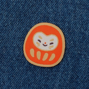 May include: An orange Daruma doll patch with a smiling face and tan accents, set against a blue denim background. The patch is oval-shaped and features a traditional Japanese design, perfect for adding to clothing or accessories.