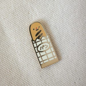 Peut inclure: Une broche en émail et métal doré représentant une baguette souriante de couleur beige avec un petit point rouge en guise de nez. La baguette est partiellement enveloppée dans un emballage blanc quadrillé avec une étiquette ovale portant le texte japonais パン.