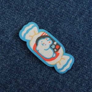Op de afbeelding: Een kleurrijke patch in de vorm van een verpakt snoepje. De patch heeft een cartoon konijn in de verpakking, met rode, witte en blauwe accenten. De patch is blauw omlijnd en geplaatst tegen een blauwe denim achtergrond.