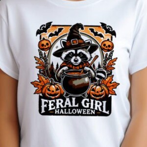 Pode incluir: Camiseta branca com um gráfico de um guaxinim usando um chapéu de bruxa e segurando um caldeirão. O gráfico é cercado por morcegos, abóboras e folhas. O texto "Feral Girl - Halloween" está abaixo do gráfico.