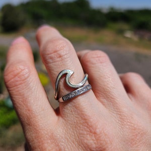 May include: A silver ring with a wave design and a row of clear stones. The ring is on a person's finger.
