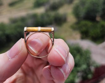 May include: A gold ring with a rectangular tiger's eye stone. The ring is held up against a blurred background of green hills and a blue sky. The ring's band is thin and delicate.