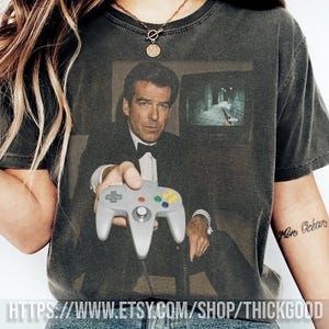 Könnte beinhalten: Dunkelgraues T-Shirt mit einem Aufdruck eines Mannes im Smoking, der einen grauen Nintendo 64-Controller hält. Das Bild auf dem Shirt enthält einen Screenshot aus einem Videospiel. Der Text "https://www.etsy.com/shop/thickgood" ist unten aufgedruckt.