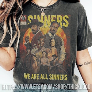 Puede incluir: Camiseta gris oscuro con un gráfico de estilo vintage. El gráfico presenta la palabra "SINNERS" en letras amarillas goteando, ilustraciones de personas y el texto "WE ARE ALL SINNERS".