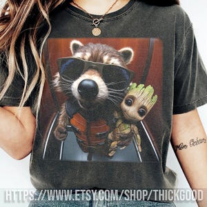 Pode incluir: Camiseta cinza escuro com um gráfico de Rocket Raccoon usando óculos de sol e Groot. A imagem é do filme Guardiões da Galáxia. O texto na parte inferior da camiseta diz HTTPS://WWW.ETSY.COM/SHOP/THICKGOOD.