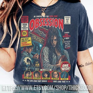 May include: A charcoal gray t-shirt featuring a graphic print designed like a vintage comic book cover titled OBSESSION. The artwork includes a central female figure, a box labeled ONE WISH WILLOW, and text including BE CAREFUL WHO YOU WISH FOR.