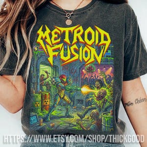 Puede incluir: Camiseta gris oscuro con el texto "METROID FUSION" en amarillo y rojo. El gráfico representa una escena de batalla futurista con un personaje con armadura disparando a criaturas zombi. La camiseta tiene un aspecto vintage y desgastado.