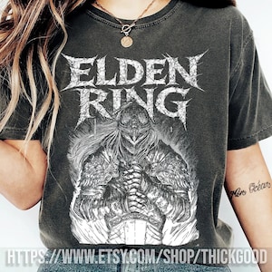 May include: Dark gray t-shirt featuring the text "ELDEN RING" in a distressed, spiky font above a detailed black and white illustration of a knight. The shirt has a vintage, worn look.