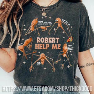 Könnte beinhalten: Dunkelgraues T-Shirt mit einem humorvollen Grafikdesign. Das Design zeigt den Text "ROBERT HELP ME" und mehrere cartoonartige, insektenähnliche Figuren. Die Grafik enthält Sätze wie "Hast du den Mondsaft probiert?" und "Ich hoffe, sie haben Tortilla-Chips."