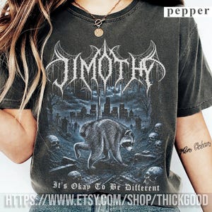 Pode incluir: Uma t-shirt cinzento-carvão de manga curta com uma impressão gráfica de um guaxinim num cemitério com o horizonte de uma cidade ao fundo. O texto "JIMOTHY" aparece numa fonte estilizada acima, com "It's Okay To Be Different" impresso abaixo.