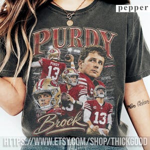 Puede incluir: Una camiseta gris carbón de manga corta con un estampado gráfico de estilo vintage del jugador de fútbol americano Brock Purdy con el uniforme de los San Francisco 49ers. El diseño incluye varias imágenes de acción y el nombre Brock.