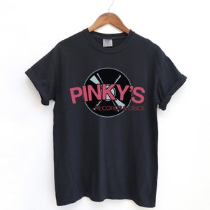 May include: Black t-shirt with a pink logo that says "Pinky's Records & Discs". The logo features a stylized record with crossed needles.