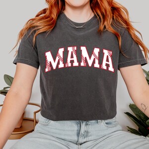 May include: A dark gray t-shirt with a white graphic of the word "MAMA" in a baseball font with red stitching.