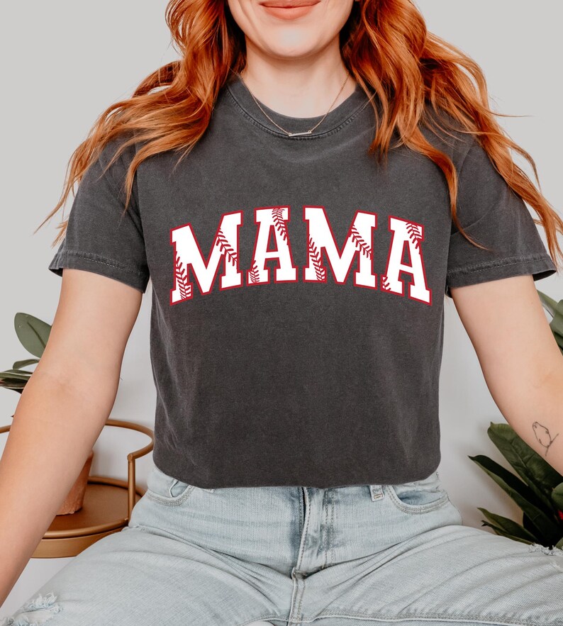 May include: A dark gray t-shirt with a white graphic of the word "MAMA" in a baseball font with red stitching.
