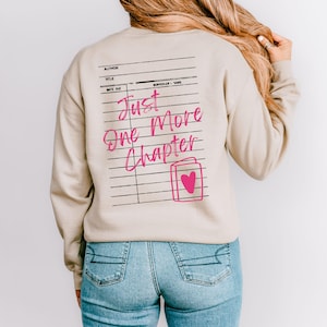 May include: A beige sweatshirt with a pink graphic of a library card. The graphic says "Just One More Chapter" and features a pink heart on a book.