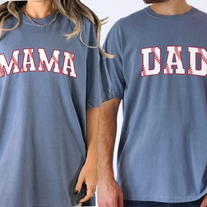 May include: Two blue t-shirts with white text and red baseball stitching. The text on the left shirt says "MAMA" and the text on the right shirt says "DAD".