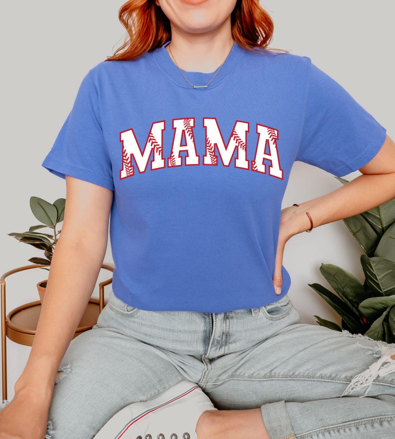May include: A royal blue t-shirt with a white baseball-themed "MAMA" graphic. The letters are outlined in red and have a baseball stitch design.