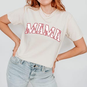 May include: A white t-shirt with the word "MAMA" printed in red with a baseball stitch design.