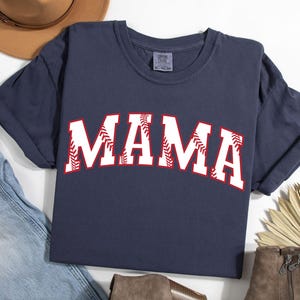 May include: A navy blue t-shirt with the word "MAMA" in white letters outlined in red. The letters are decorated with a baseball stitch design.