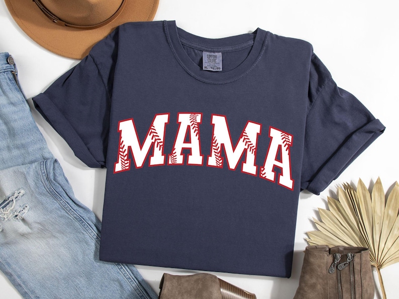 May include: A navy blue t-shirt with the word "MAMA" in white letters outlined in red. The letters are decorated with a baseball stitch design.