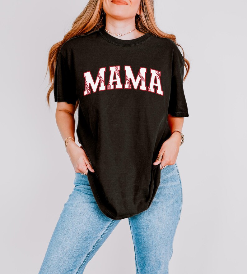 May include: A black t-shirt with the word "MAMA" in white letters outlined in red. The letters are styled to look like a baseball. The shirt is being worn by a person with long brown hair.