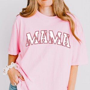 May include: Pink t-shirt with a white "MAMA" graphic. The letters are outlined in maroon and have a baseball stitch design.