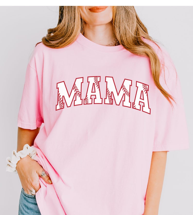 May include: Pink t-shirt with a white "MAMA" graphic. The letters are outlined in maroon and have a baseball stitch design.