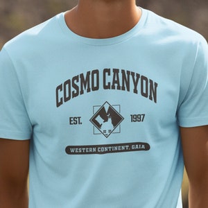 Puede incluir: Una camiseta azul claro con un estampado gráfico marrón que dice "COSMO CANYON EST. 1997 WESTERN CONTINENT. GAIA".