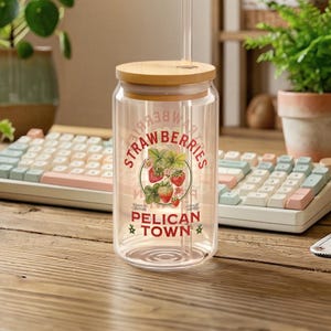 Cozy desk scene featuring a Stardew Valley inspired glass cup with a bamboo lid and straw, decorated with "Strawberries Pelican Town" artwork, set on a wooden table with a pastel mechanical keyboard, houseplants, and warm ambient lighting.