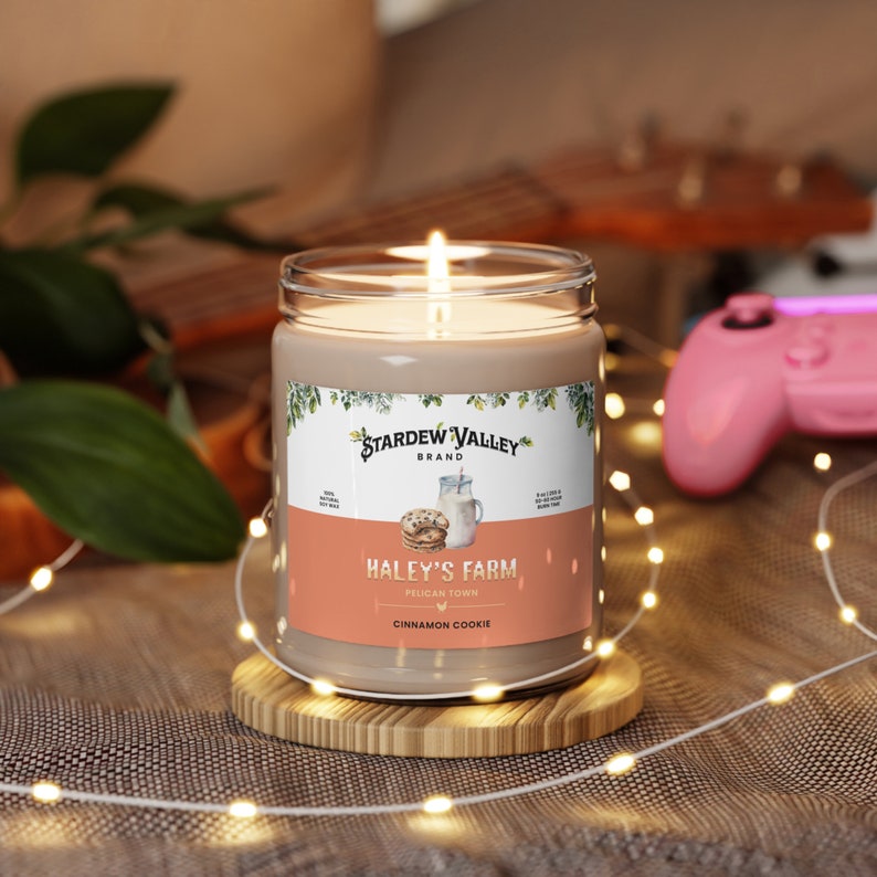 May include: A lit candle in a glass jar with a label that reads "Haley's Farm, Pelican Town, Cinnamon Cookie." The candle sits on a wooden coaster. The background includes a guitar, a pink game controller, and string lights.