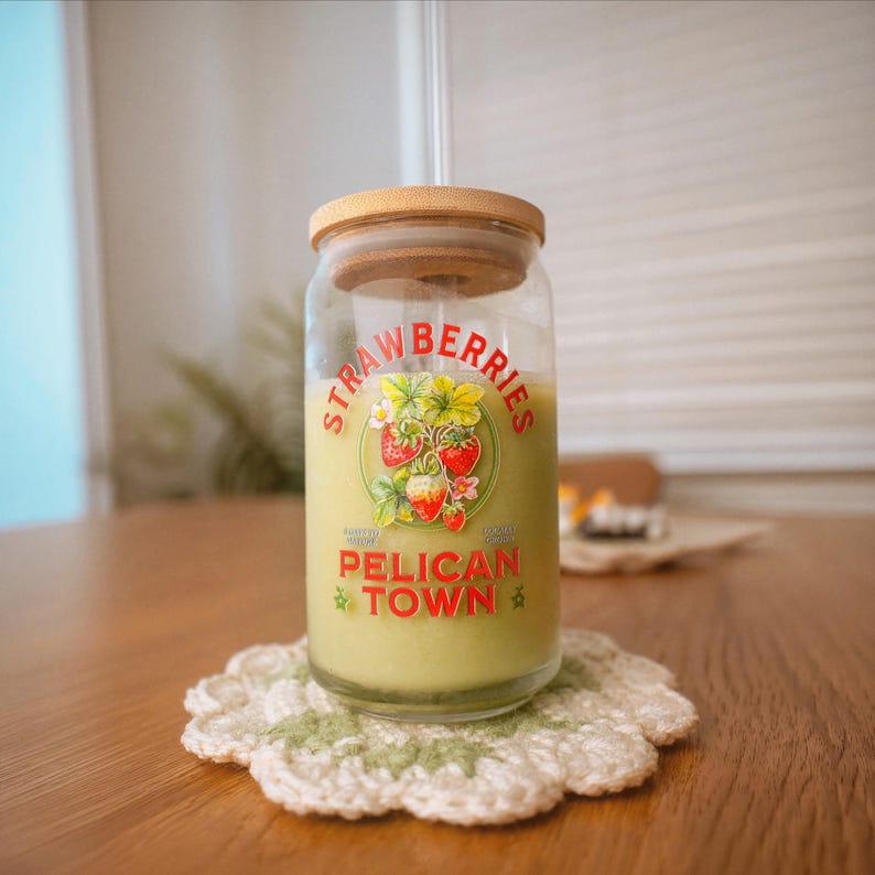 Op de afbeelding: Een glazen pot met een houten deksel, gevuld met een groene vloeistof en een aardbeiengrafiek. De pot staat op een wit en groen gehaakt onderzetter. De tekst op de pot luidt "Strawberries" en "Pelican Town."