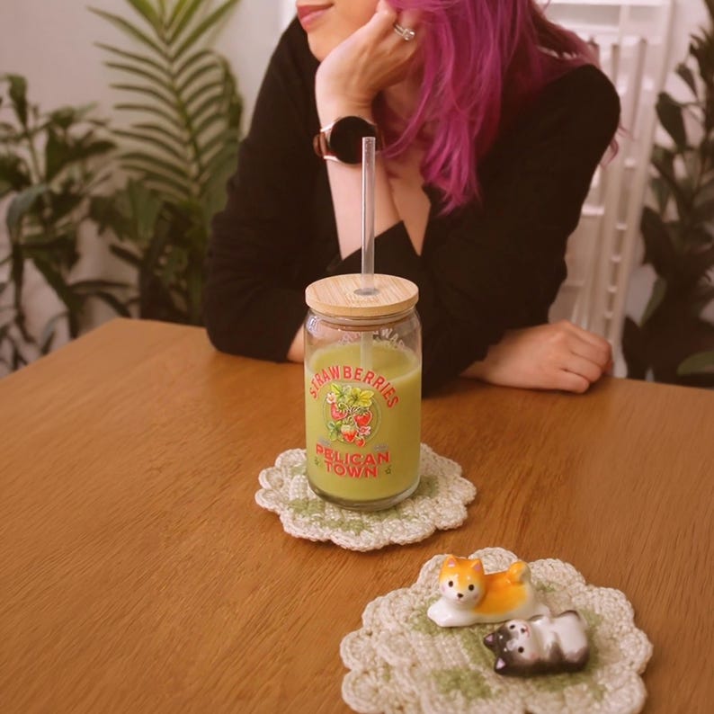 Op de afbeelding: Een glazen pot met een houten deksel en een rietje, gevuld met een groene drank, staat op een wit en groen gehaakte onderzetter. Op de pot staat "Strawberries Pelican Town". Een kleine keramische vos en hond staan op een vergelijkbare onderzetter.