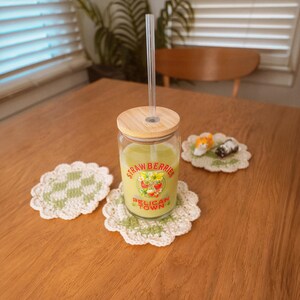 Op de afbeelding: Een glazen beker met een houten deksel en een doorzichtig rietje, gevuld met een groene drank, staat op een gehaakte onderzetter. De beker heeft een "Strawberries Pelican Town" label. Twee andere gehaakte onderzetters en een klein decoratief item liggen op de houten tafel.