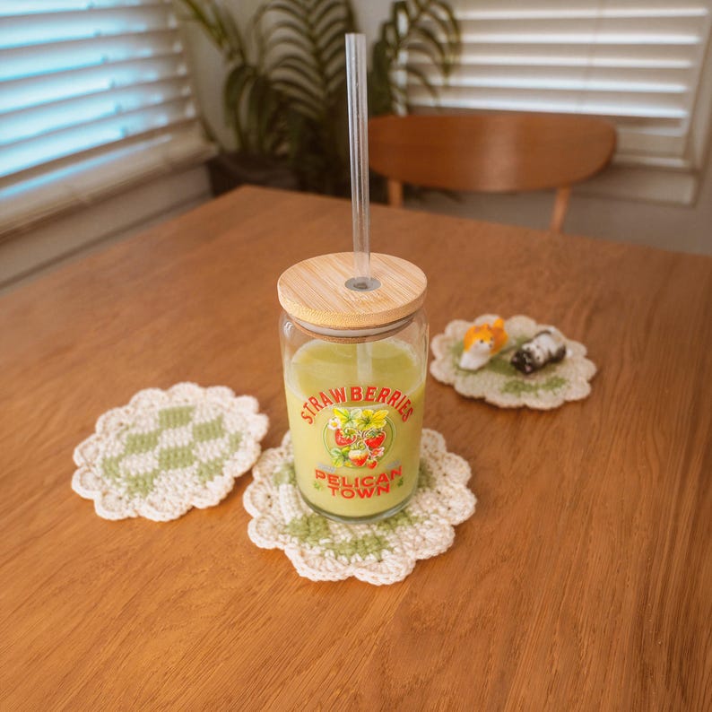 Op de afbeelding: Een glazen beker met een houten deksel en een doorzichtig rietje, gevuld met een groene drank, staat op een gehaakte onderzetter. De beker heeft een "Strawberries Pelican Town" label. Twee andere gehaakte onderzetters en een klein decoratief item liggen op de houten tafel.
