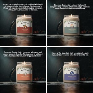 May include: Four glass jar candles with lit wicks. Each candle has a label with the "Stardew Valley Brand" logo and a unique scent name: Apple Cider, Cindersap Blooms, Cinnamon Cookie, and Dance of the Moonlight. The candles are on a dark surface.