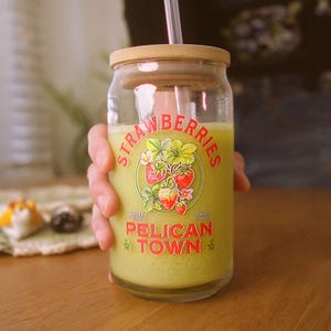 Op de afbeelding: Een heldere glazen pot met een bamboe deksel en rietje, gevuld met een groene drank. De pot heeft een vintage label met het woord "STRAWBERRIES" en de tekst "PELICAN TOWN" met aardbeien illustraties.