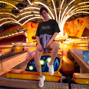 A woman at night sitting on a carnival ride wearing a black Cyberpunk 2077 Hey Choom Unisex Oversized T-Shirt by New Save.