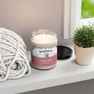 May include: A lit candle in a clear glass jar with a label that reads "Leah's Farm" and "Sea Salt Sweet Pea." The candle sits on a white surface next to a ball of white yarn and a small potted plant. The candle's label also says "Stardew Valley Brand."