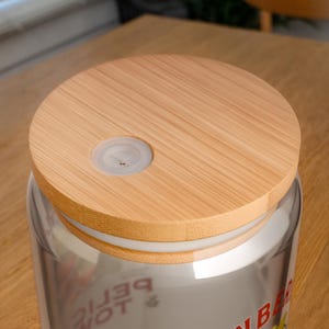 Op de afbeelding: Een heldere glazen pot met een lichtbruine bamboe deksel. De deksel heeft een kleine, cirkelvormige, doorzichtige plastic inzet. De pot staat op een lichtbruin houten oppervlak. De pot lijkt een drankje te bevatten.